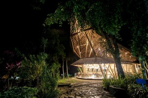 Una estructura de bambú con una zona de estar circular, rodeada de exuberante vegetación y un sendero de piedra.