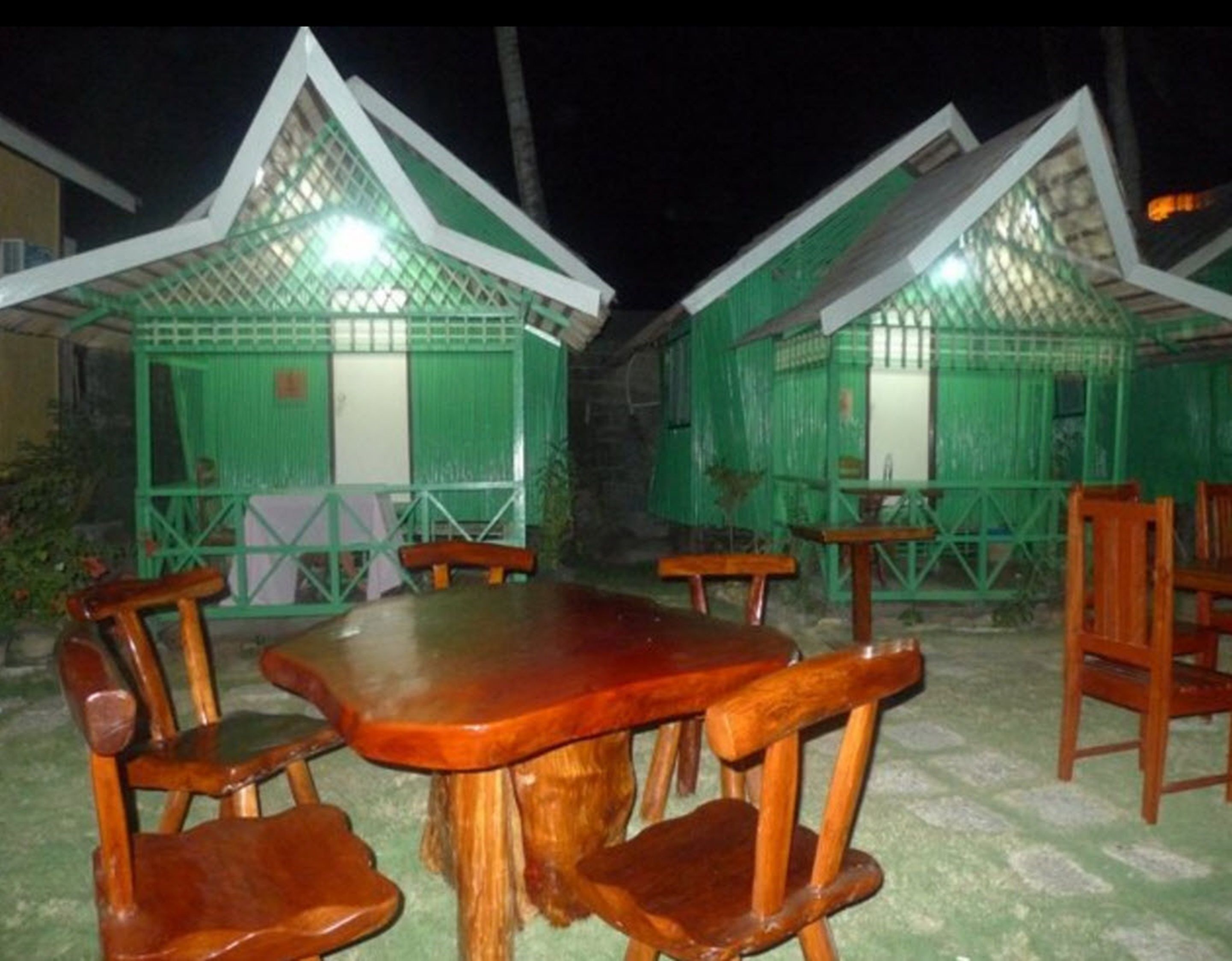 A row of green-roofed cottages with wooden furniture, including a table and chairs, set on a stone-paved ground.