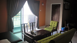 A living room with a green sofa, a wooden coffee table, and a green armchair. There is a window with curtains, a door, and a small table with a lamp. - Aeon Tebrau Apartment Johor Bahru (Johor Bahru)