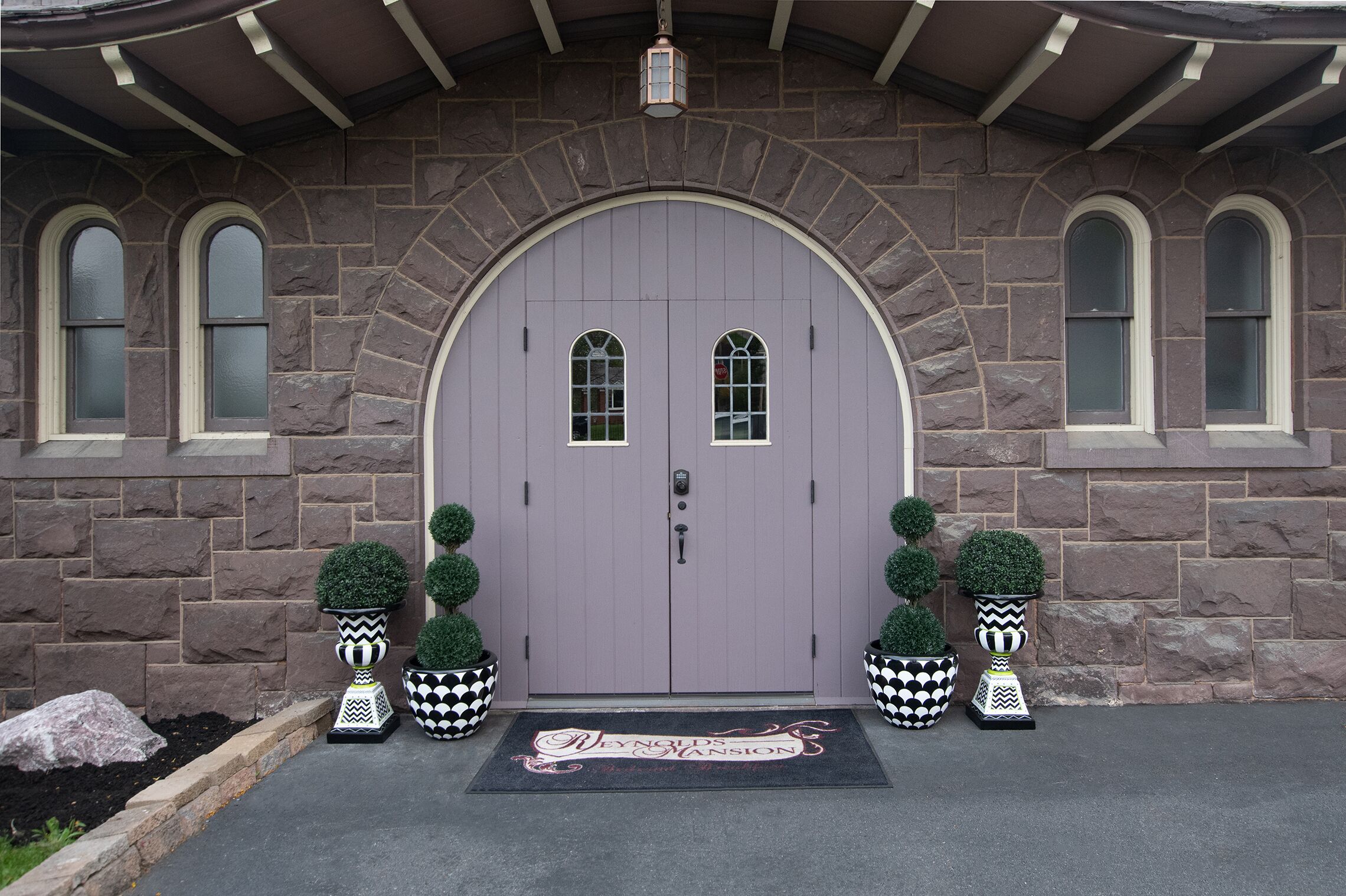 A stone building with a large arched entrance door, two windows on either side, and a doormat with the name "Reynolds Mansion".