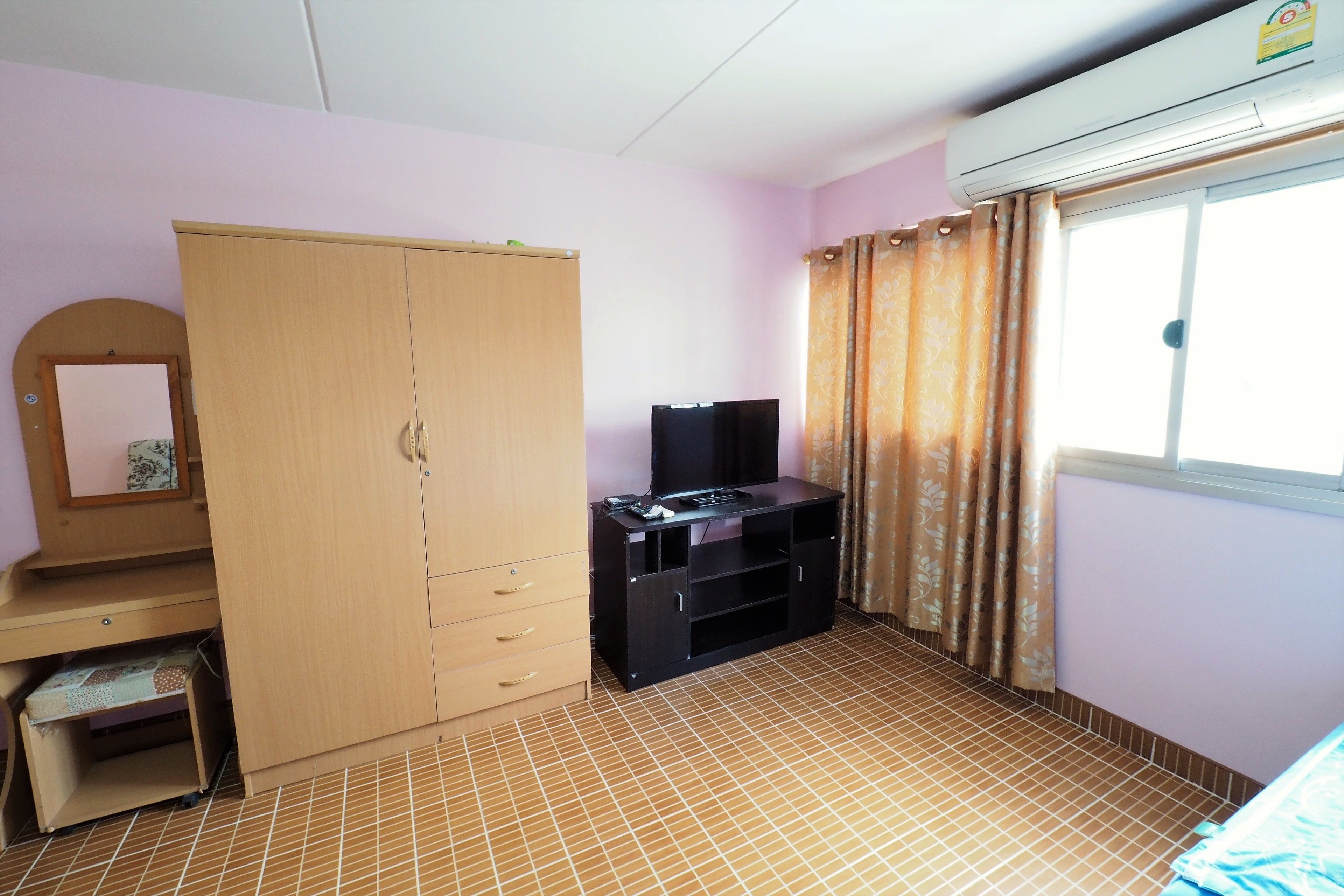 A room with a wardrobe, a television on a stand, a window with curtains, and a tiled floor.