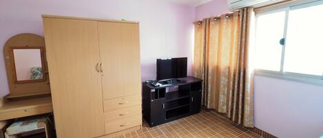 A room with a wardrobe, a television on a stand, a window with curtains, and a tiled floor.