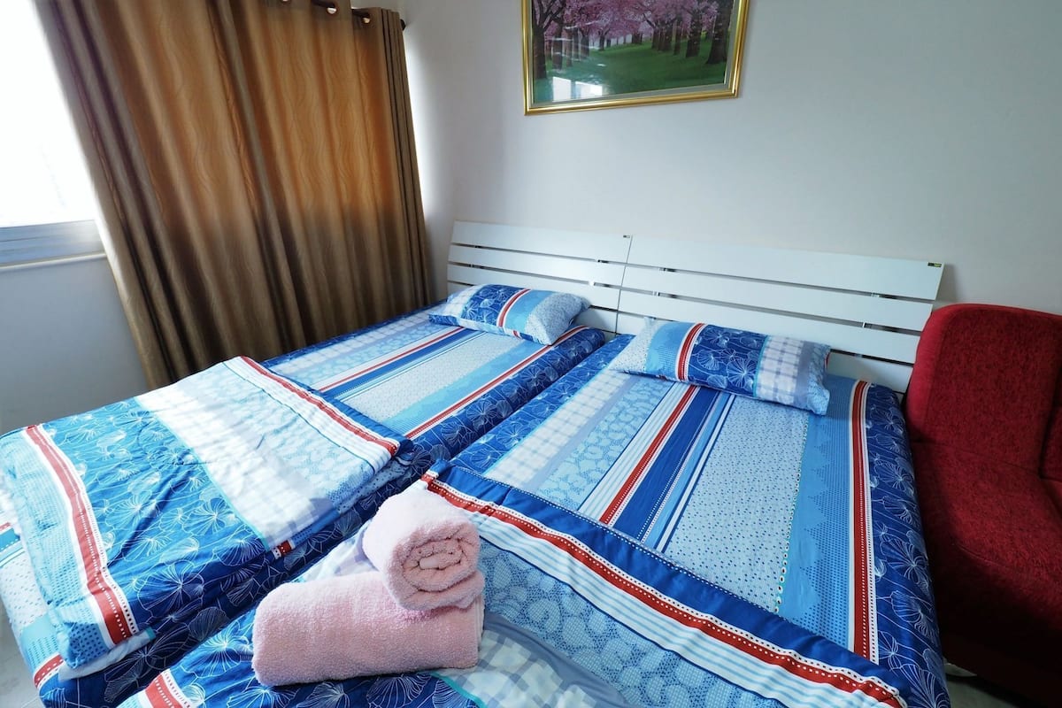 Ein ordentlich bezogenes Bett mit einem gemusterten Überwurf, ein aufgerolltes Handtuch und ein roter Sessel daneben.