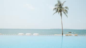 A clear blue infinity pool with white lounge chairs and a palm tree, overlooking the sea.