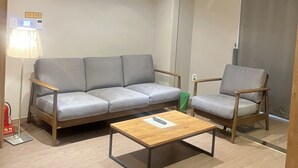 A modern living room with a grey sofa, a wooden coffee table, and a wooden armchair. There is a floor lamp and a green trash bin visible.