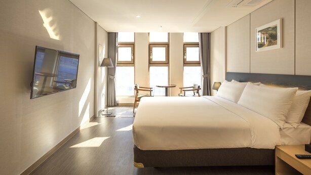 A hotel room with a large bed, a TV mounted on the wall, a desk with a lamp, and a view of the city through large windows.