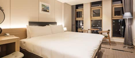 A hotel room with a large bed, a desk with a telephone, a chair, a small table, and a window with curtains.