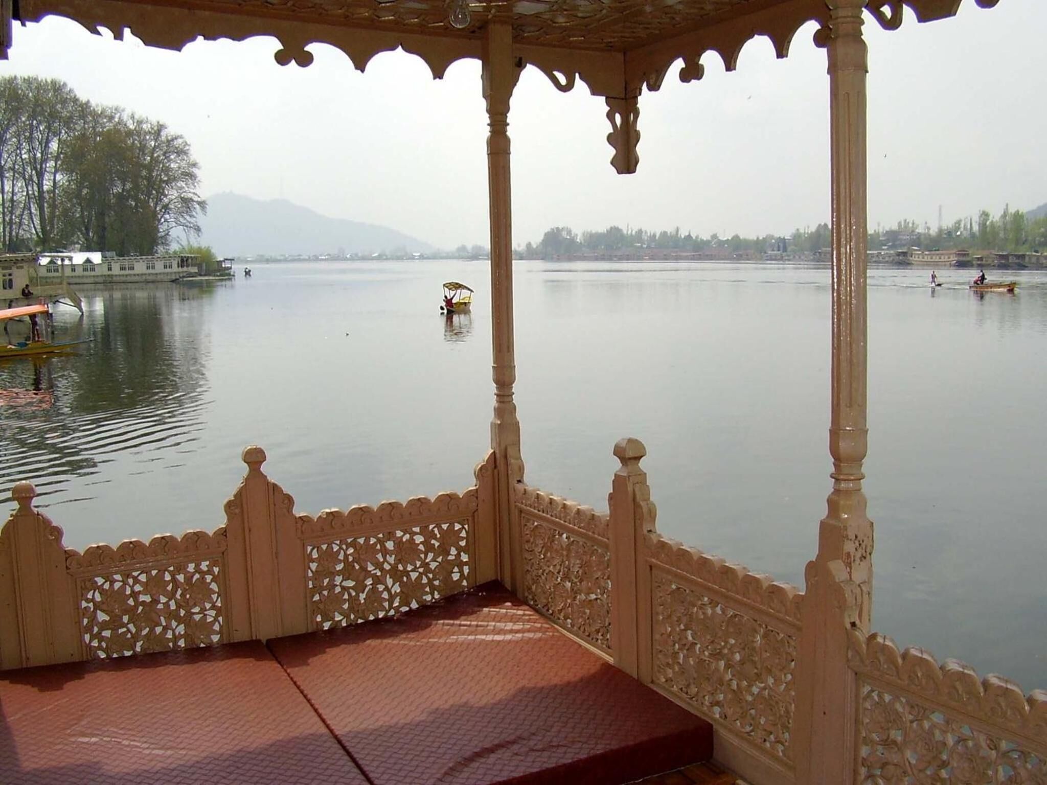 A gazebo with intricate carvings, overlooking a calm lake with boats and distant buildings.