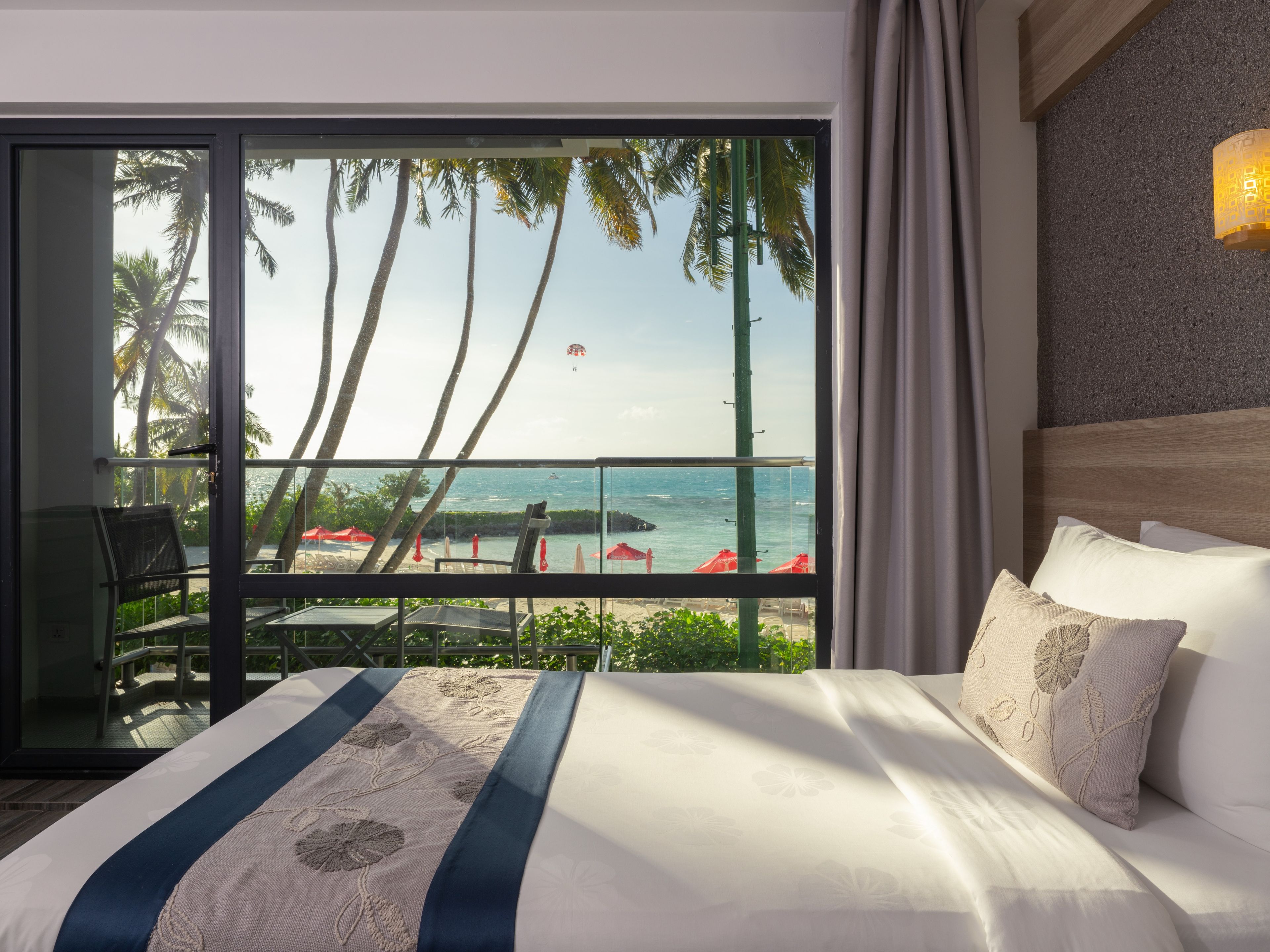 Una habitación de hotel con una cama grande, vista al océano y un balcón con sombrillas rojas.