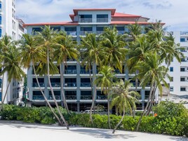 Un hotel de varios pisos con balcones, rodeado de palmeras y un primer plano con arena.