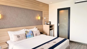 A hotel room with a large bed, a wooden headboard, a wall-mounted light, a small desk with a kettle, and a door with a frosted glass panel. - Kaani Grand Seaview (Maafushi)