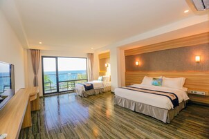 Quarto de hotel com duas camas, cabeceira de madeira, uma janela grande com vista para o oceano e uma TV de tela plana sobre uma escrivaninha de madeira.