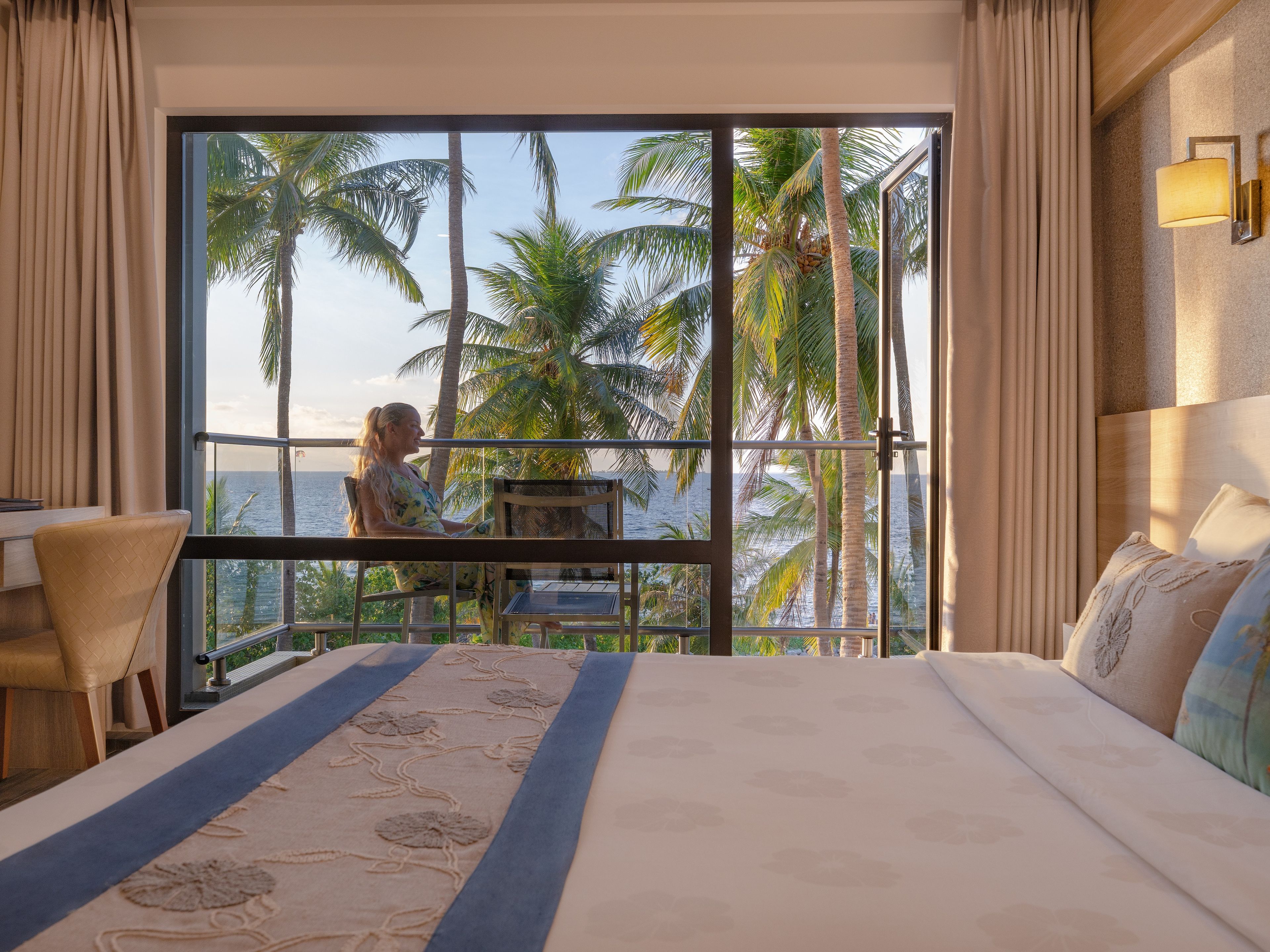 A hotel room with a large bed, a balcony with a view of palm trees and the sea, and a desk with a chair.