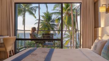 A hotel room with a large bed, a balcony with a view of palm trees and the sea, and a desk with a chair.