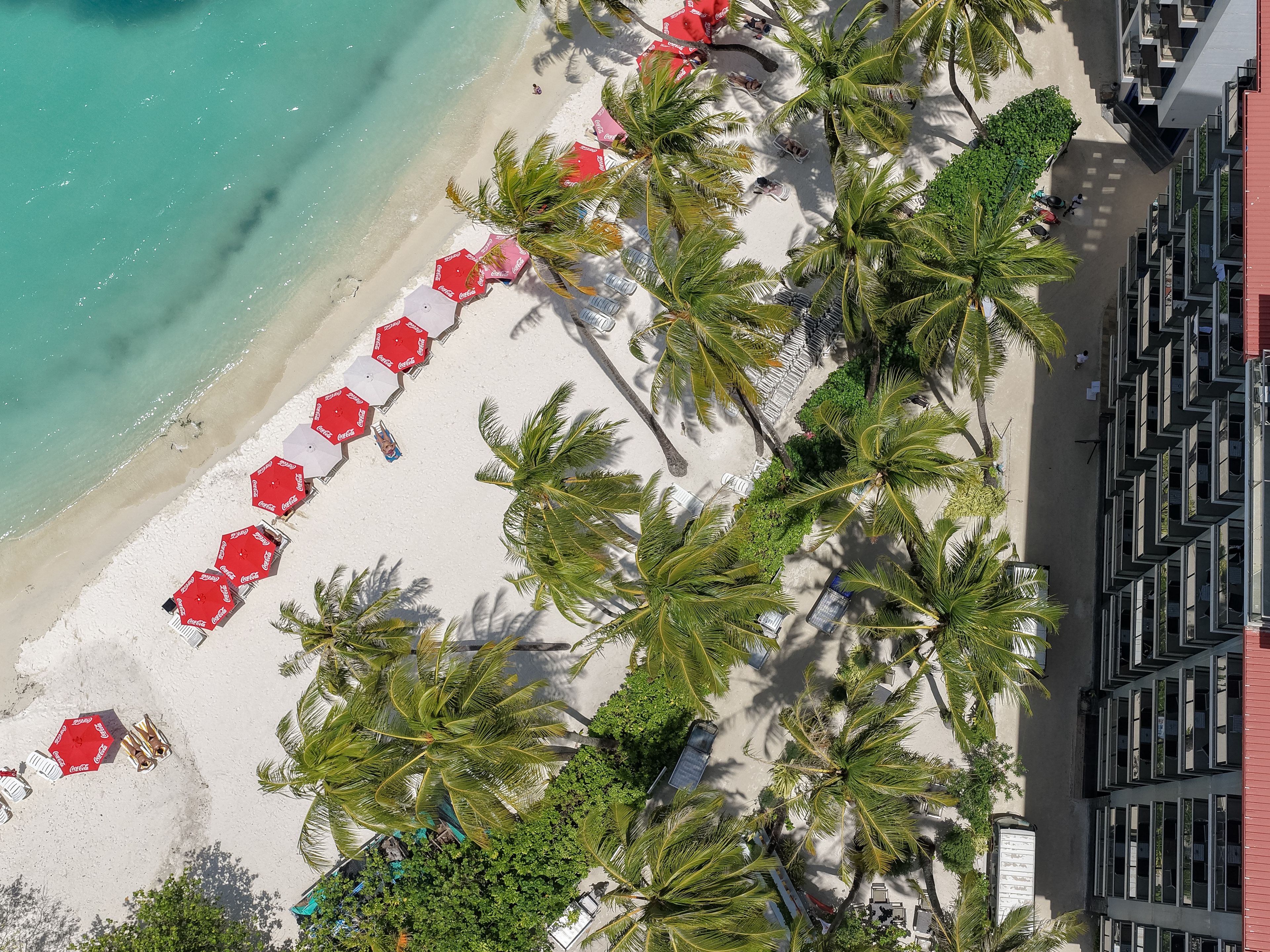 An aerial view of a beach with rows of red umbrellas, palm trees, and a building complex.