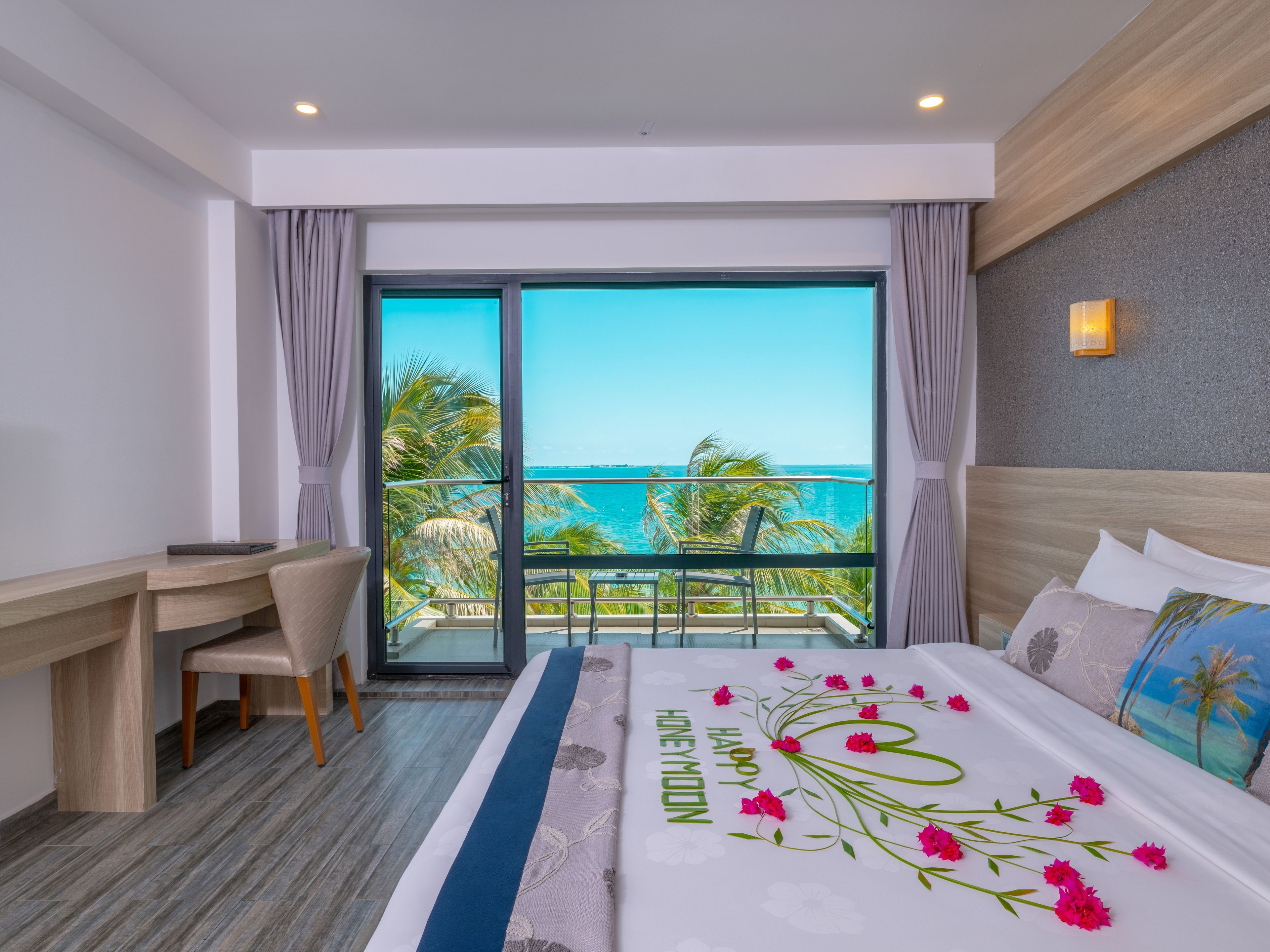 A hotel room with a bed, a desk, a chair, a balcony with a view of the ocean, and a large window.