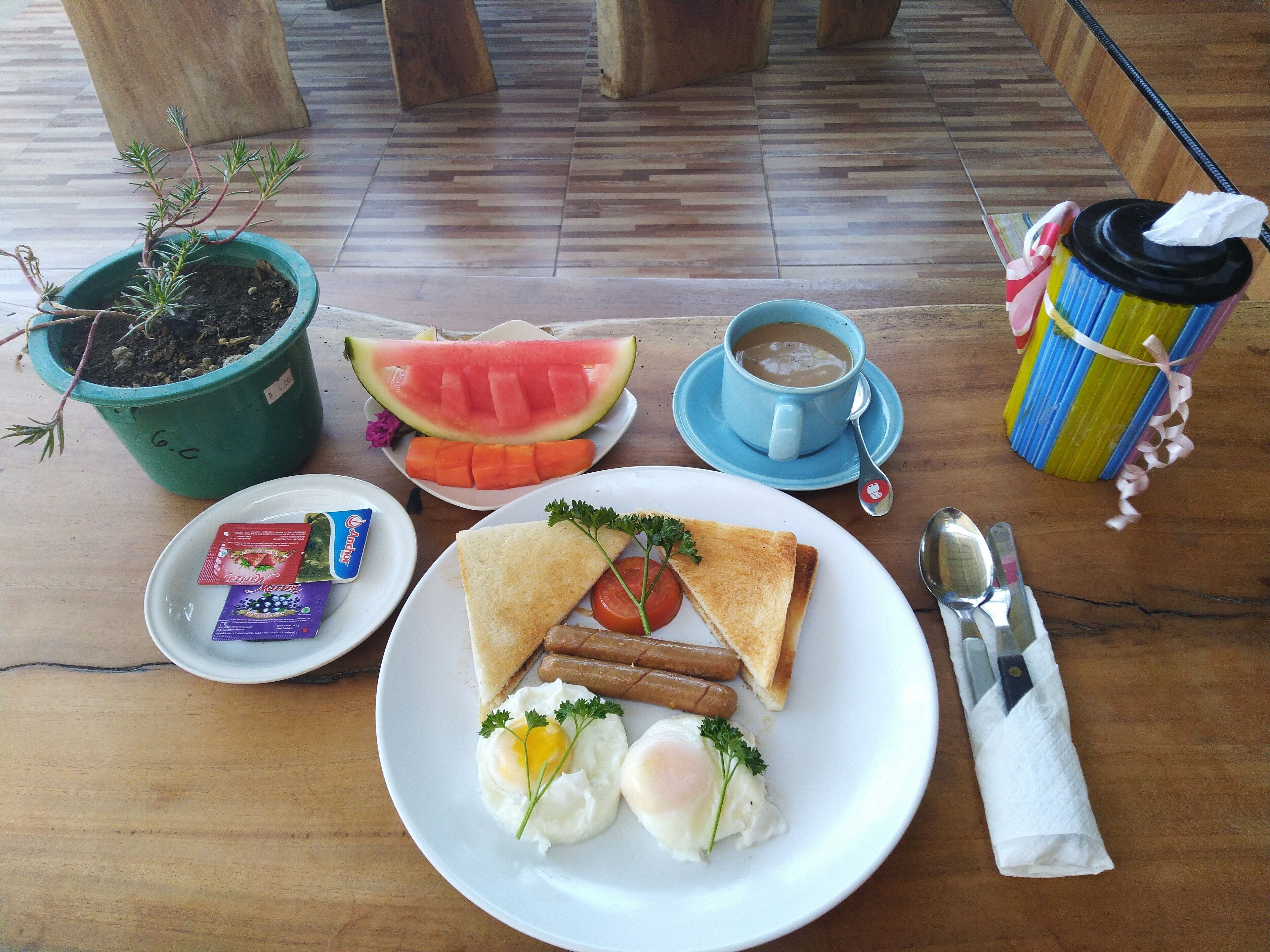 A breakfast setting with a plate of eggs, toast, sausages, and a slice of watermelon, a cup of coffee, and a colorful tumbler.