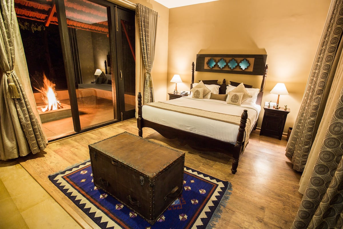 A bedroom with a bed, bedside lamps, a wooden nightstand, a suitcase, and a view of a fire outside.