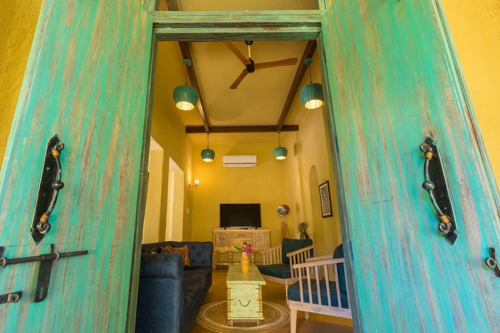 Una habitación con una puerta pintada de color verde azulado, un ventilador de techo, un televisor, una mesita con un jarrón y un sofá.