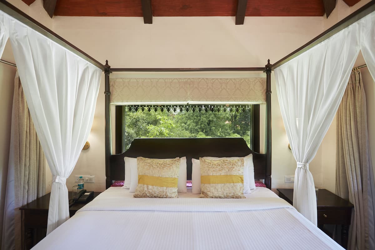 A bedroom with a large bed, white curtains, and a view of greenery outside.