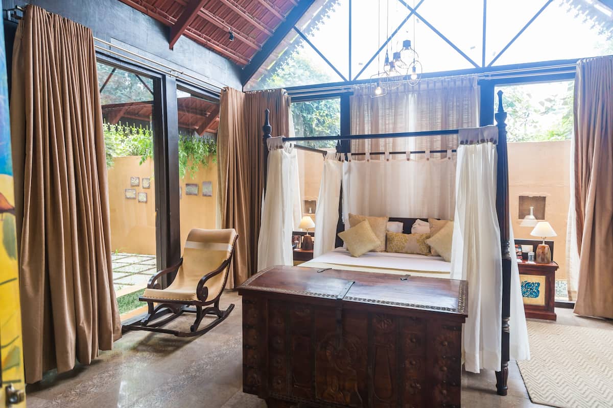 A four-poster bed with a canopy, a rocking chair, and a wooden chest of drawers in a room with large windows and curtains.