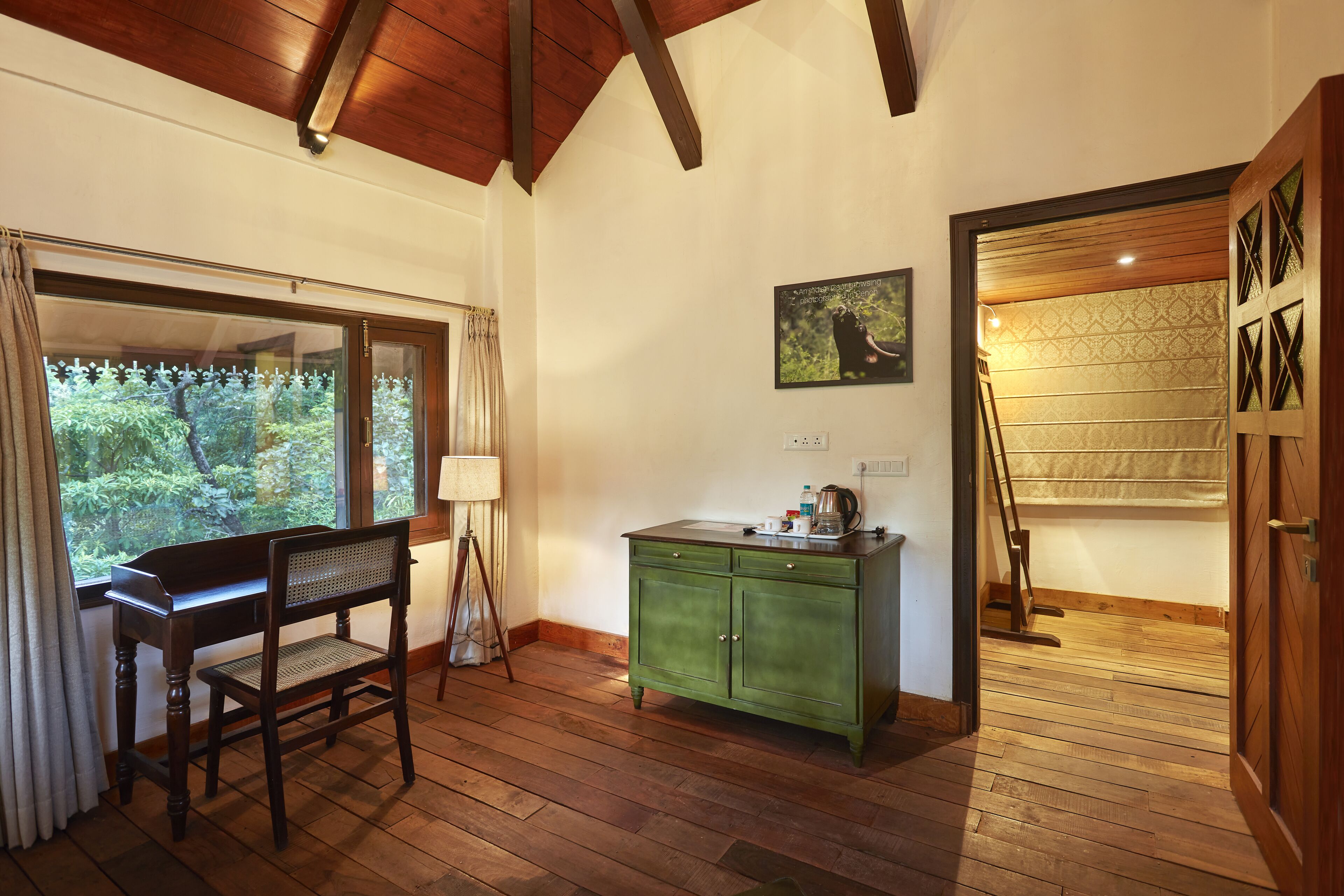 A room with a wooden floor, a green cabinet, a desk with a chair, a window with curtains, and a painting on the wall.