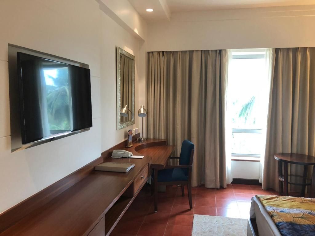 A hotel room with a flat-screen TV mounted on the wall, a desk with a telephone, a chair, a bed with a patterned bedspread, and a window with curtains.