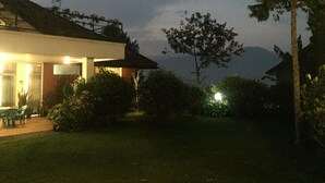 A house with a well-lit entrance, a lawn, and a mountain in the background. - Vila Bunga Merah Gelatik (Lembang)