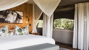 A bedroom with a canopy bed, a large framed picture, a bedside table with a lamp, and a view of greenery outside.