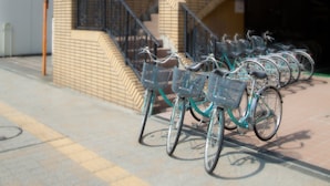 A row of bicycles with baskets parked in front of a building with a tiled facade and a staircase. - Hotel Sungarden Matsuyama (Matsuyama)