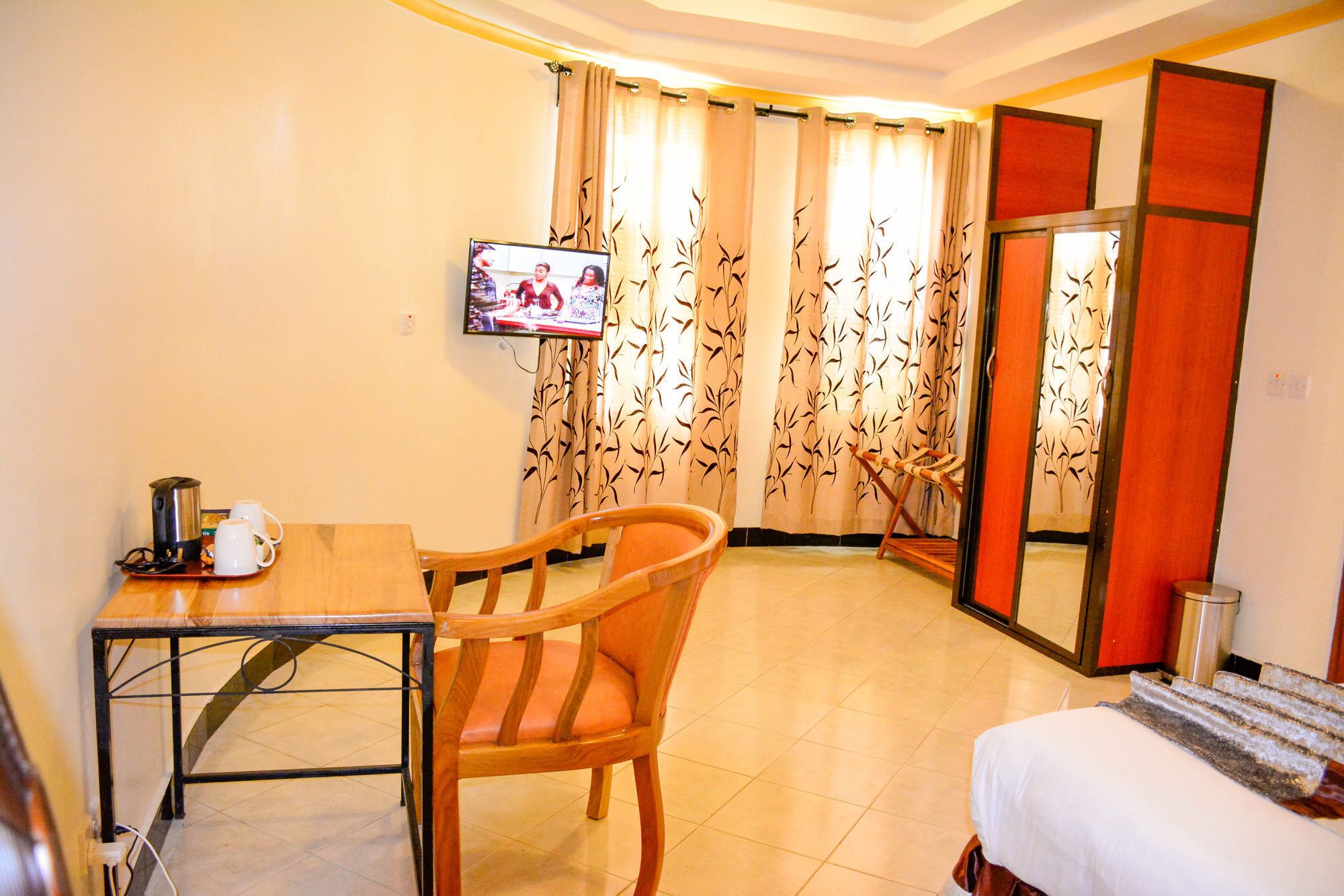 A hotel room with a bed, a wooden chair, a table with a kettle and cups, a TV on the wall, and curtains with a leaf pattern.
