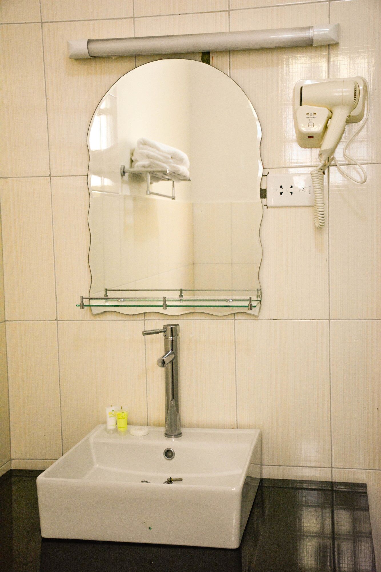 A bathroom with a rectangular sink, a mirror with a glass shelf, a towel rack, a hairdryer, and a power outlet.