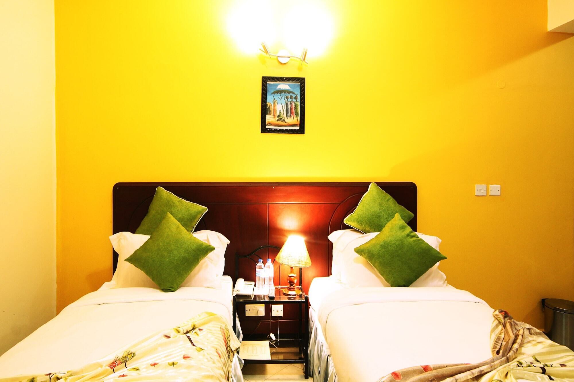 A hotel room with two beds, each with green pillows, a bedside table with a lamp, and a painting on the wall.