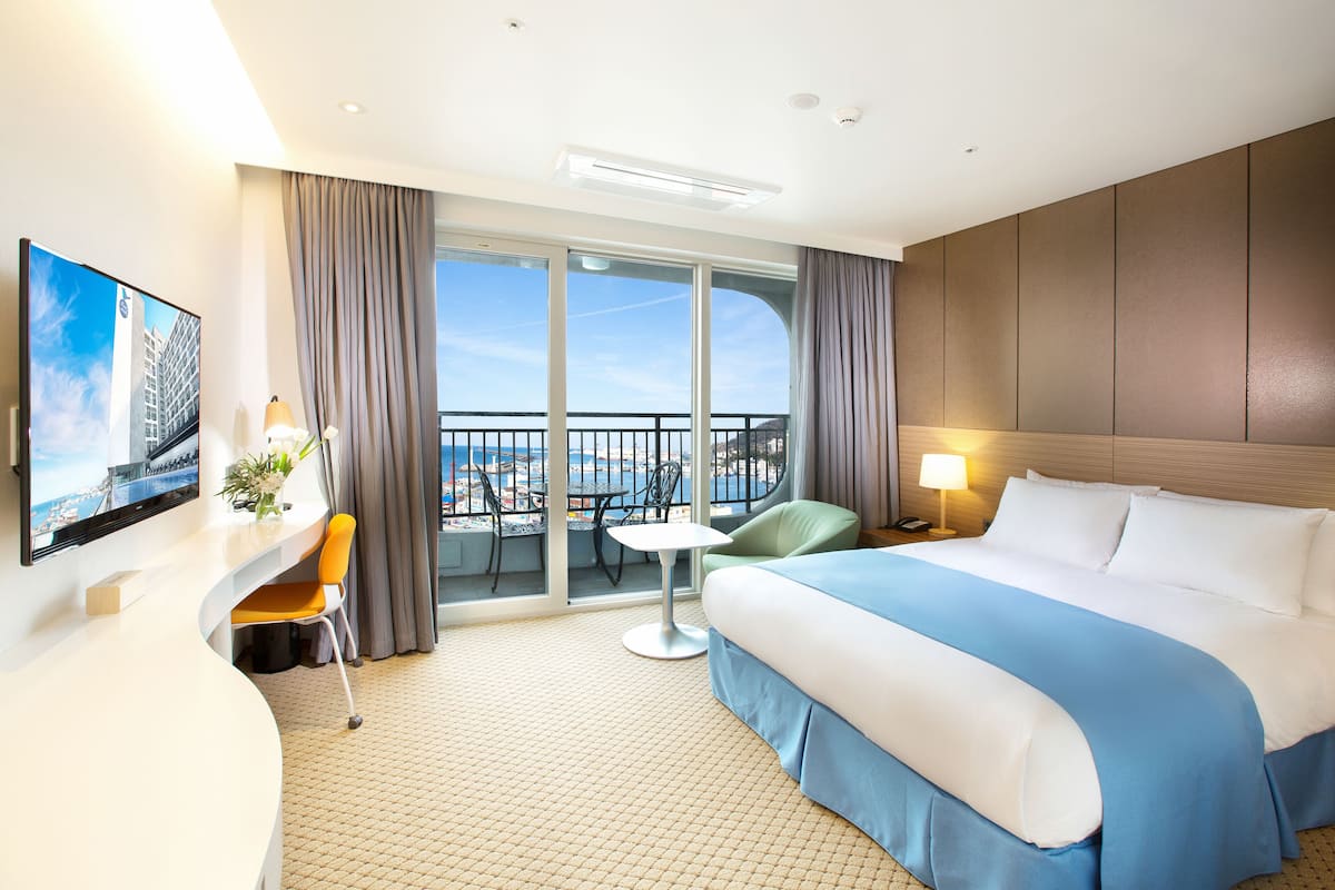 A hotel room with a large bed, a desk with a chair, a small table, and a balcony with a view of the city.