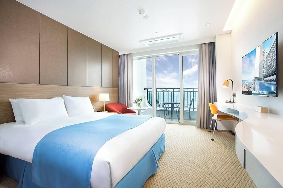 A hotel room with a large bed, a desk, a chair, a TV, and a balcony with a view.