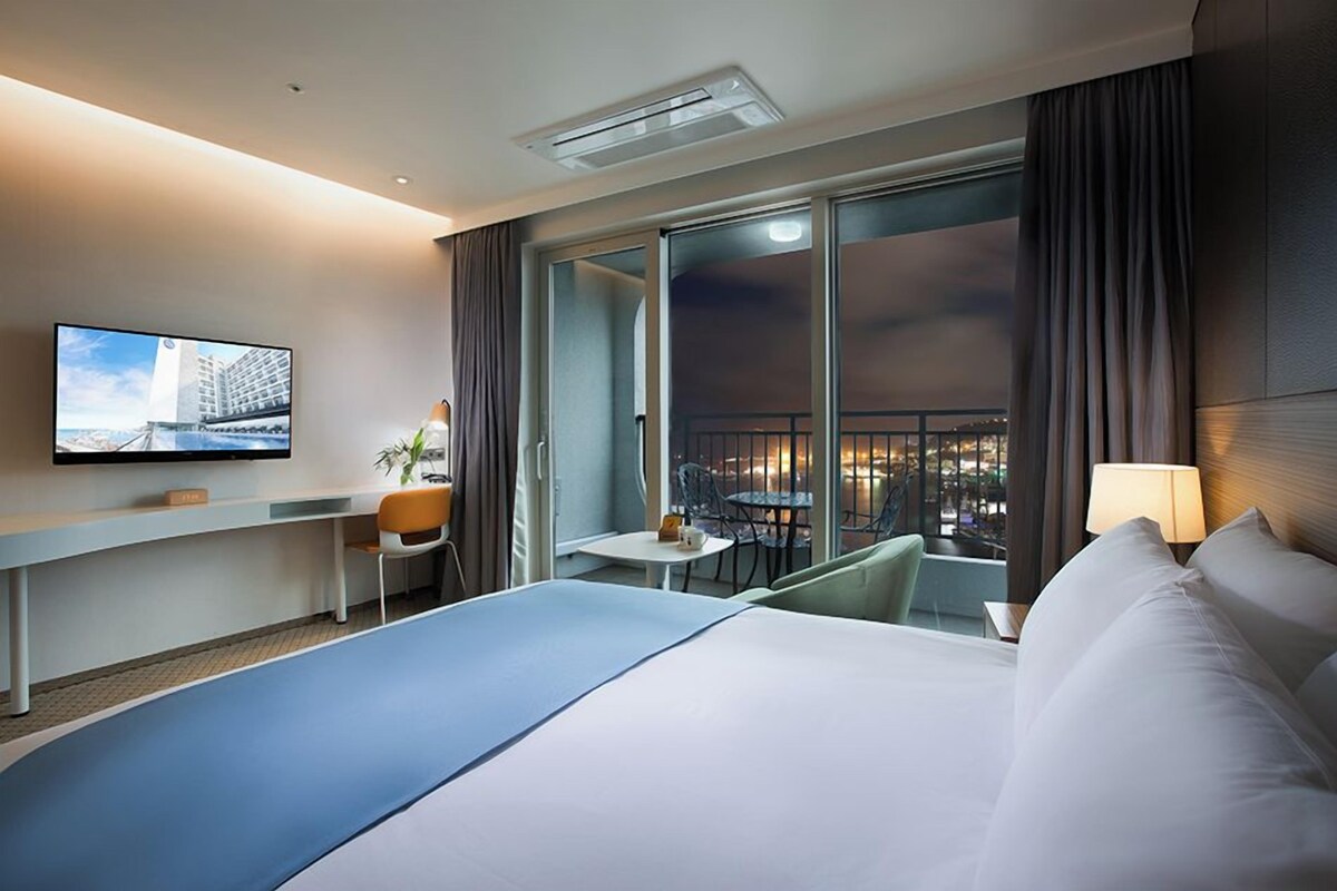 Habitación de hotel con cama, televisión, escritorio, silla, mesita y balcón con vista a la ciudad.