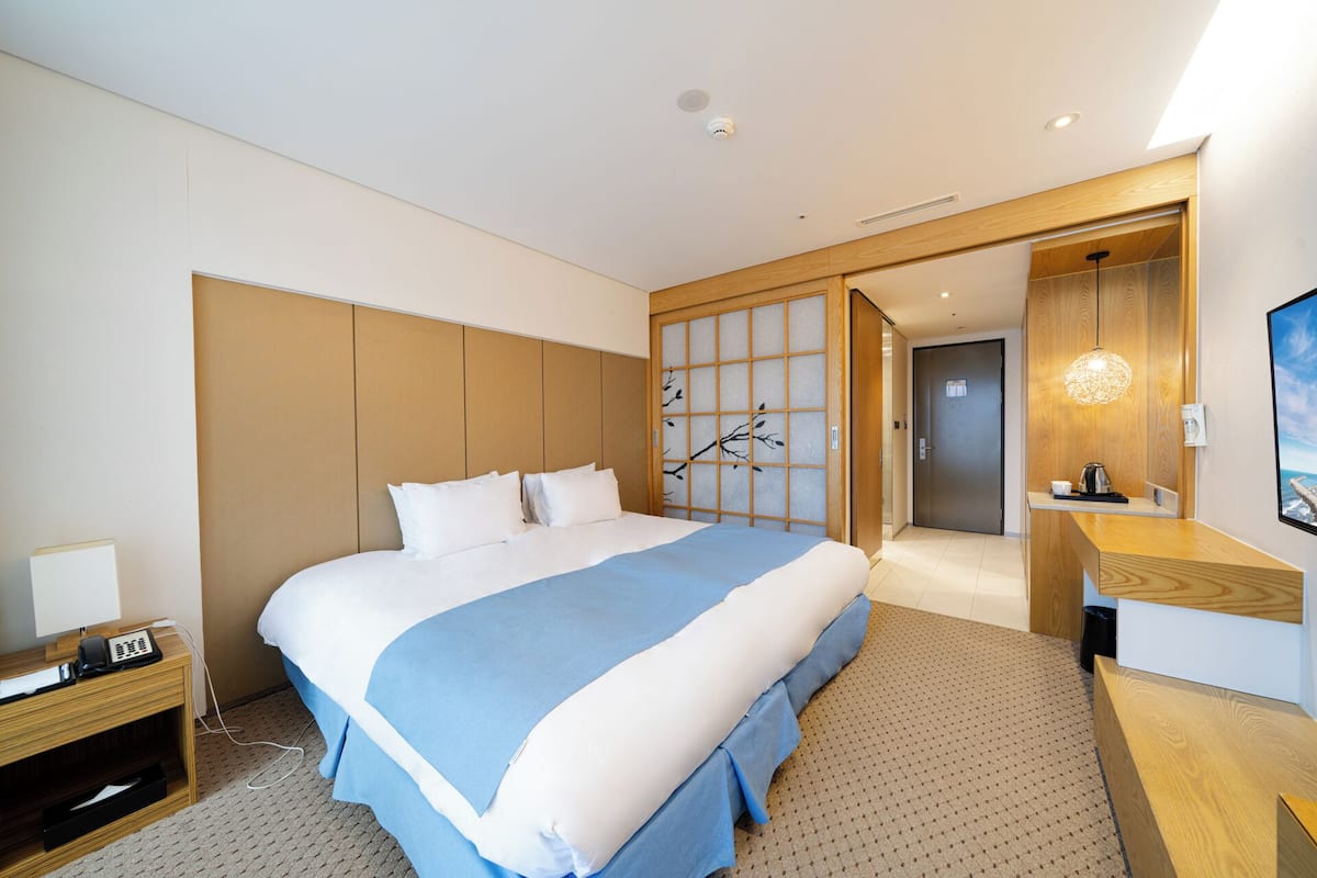 A modern hotel room with a large bed, a wooden headboard, a sliding shoji screen, a desk with a telephone, and a view of the hallway.