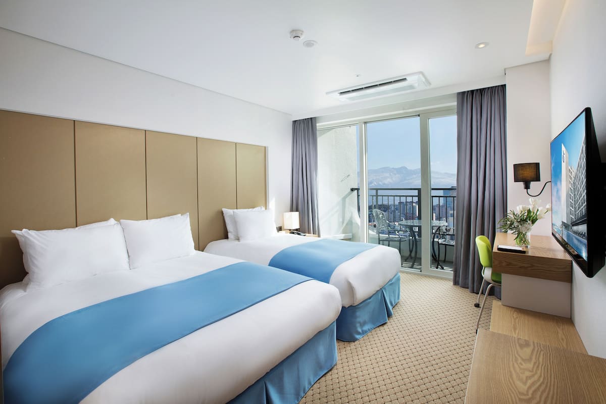 A hotel room with two beds, a desk, a chair, a TV, and a balcony with a city view.