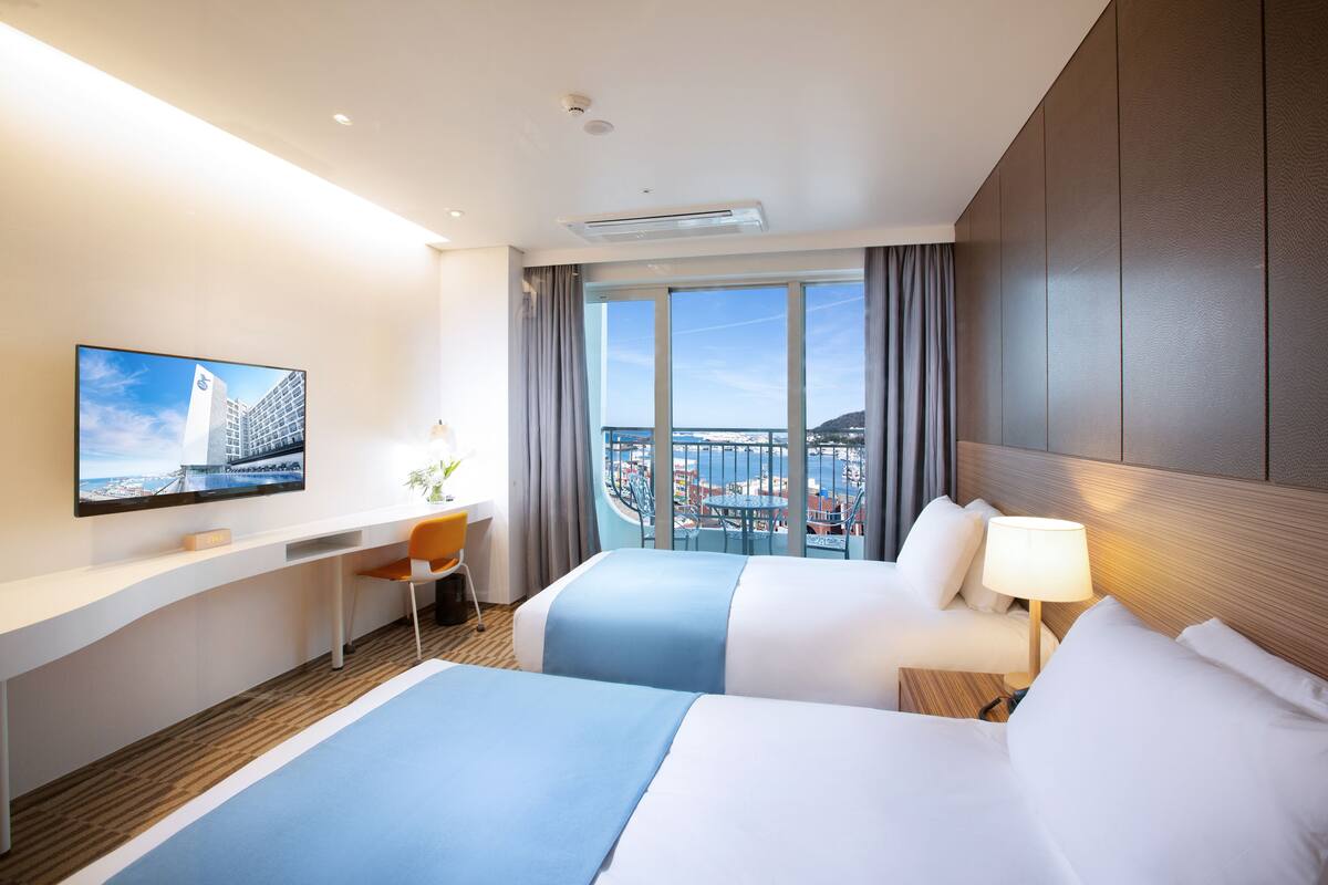 A hotel room with two beds, a desk, a chair, a TV, and a large window with a city view.