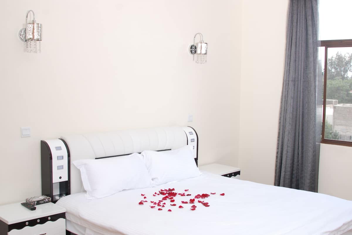 Una habitación de hotel con una cama, una mesita de noche, una lámpara de pared y un ventanal con cortinas.