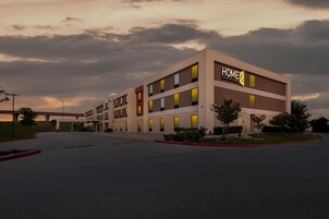 A modern hotel building with a sign reading 'HOME 2 Suites by Hilton' and a parking lot in front.