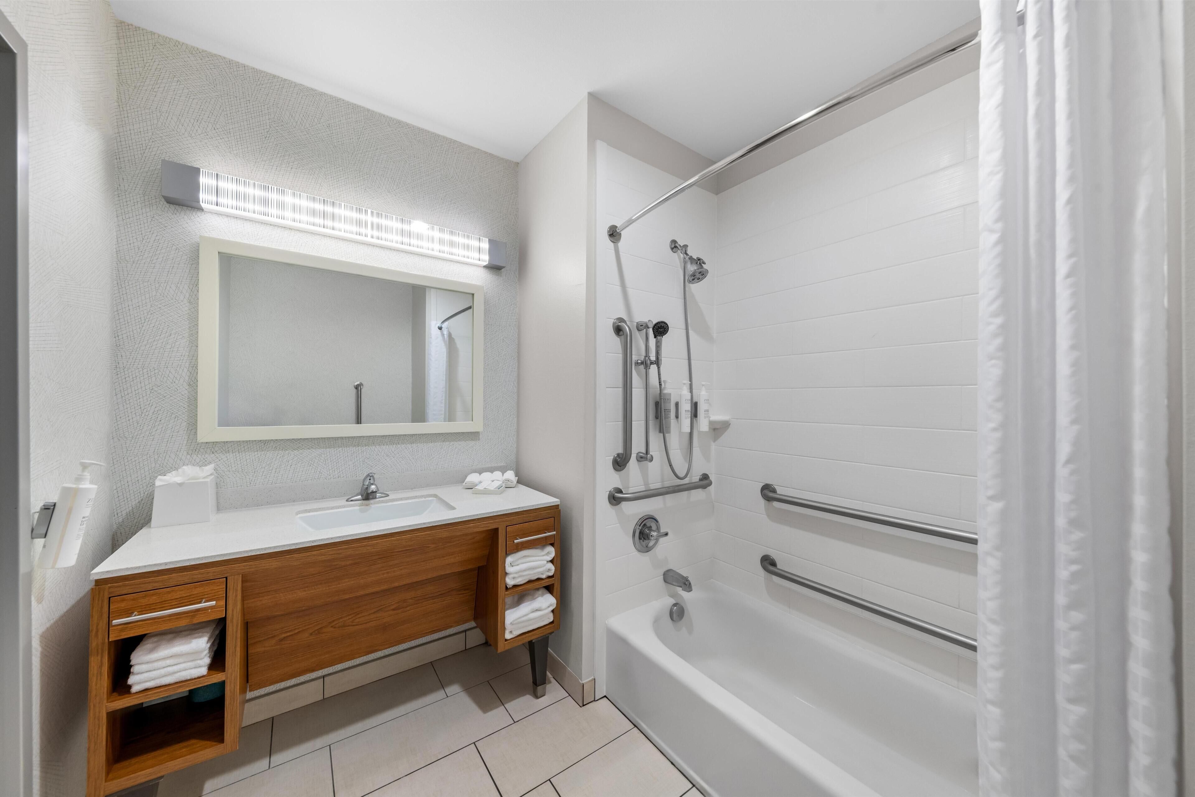 Un baño moderno con tocador doble, un espejo grande, bañera con barras de apoyo y ducha con cabezal de ducha de mano.