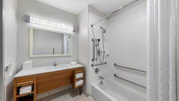 A modern bathroom with a double vanity, a large mirror, a bathtub with grab bars, and a shower with a handheld showerhead.