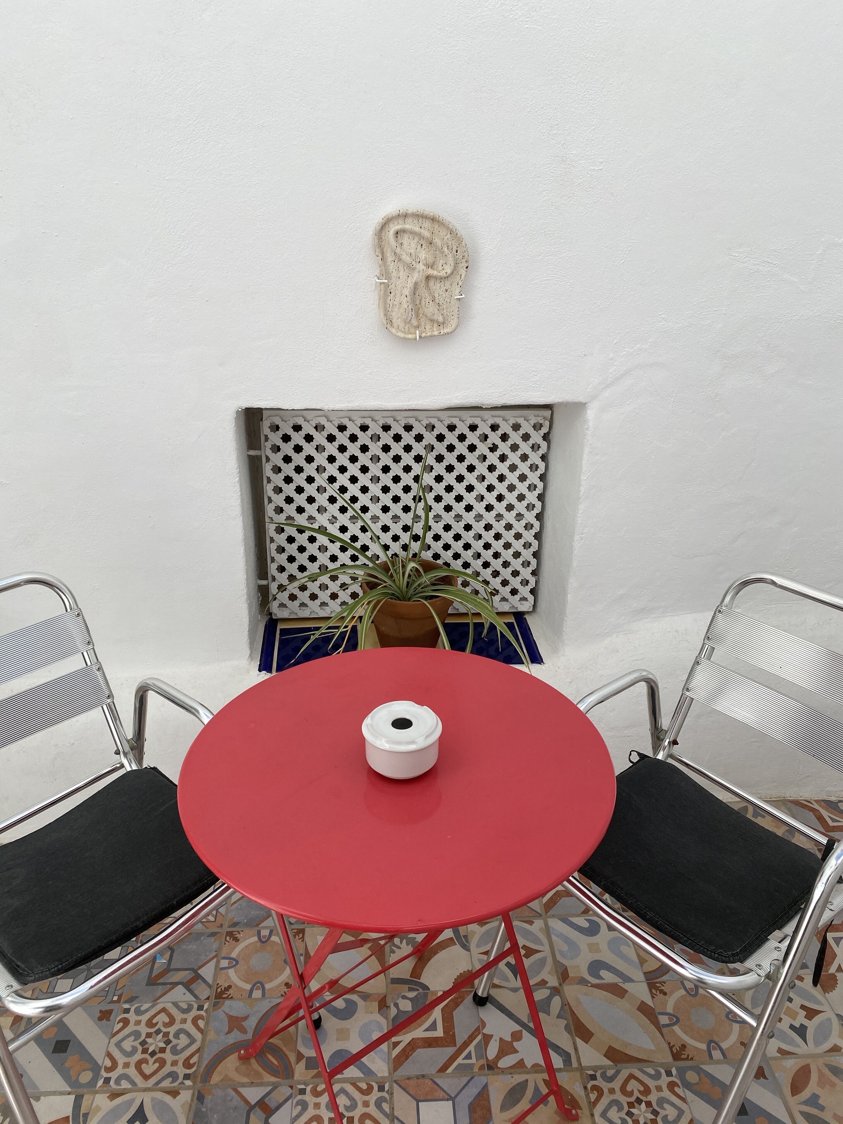 Una pequeña mesa roja con un objeto blanco encima, flanqueada por dos sillas metálicas con cojines negros. En el fondo, hay una planta en maceta y el suelo tiene baldosas con diseños.