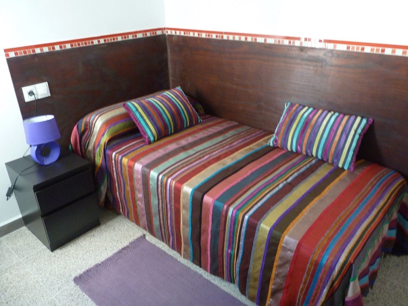 Une chambre avec une tête de lit en bois, deux lits avec une literie à rayures, une table de chevet avec une lampe et un tapis violet.
