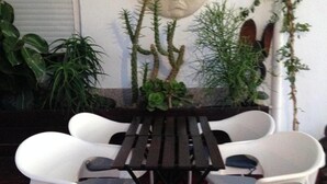 Un patio con una mesa de madera y cuatro sillas blancas de plástico. Se observa una gran escultura de piedra en la pared detrás de la mesa.