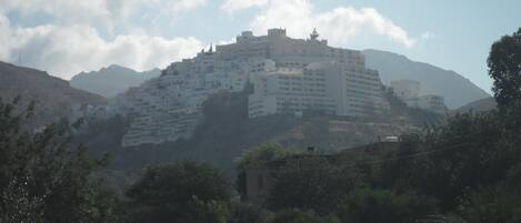 A large, multi-story building with a flat roof, situated on a hill with a mountainous backdrop.