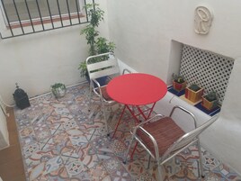 Een klein terras met een rode tafel en twee stoelen, omringd door potplanten en een patroon vloer.