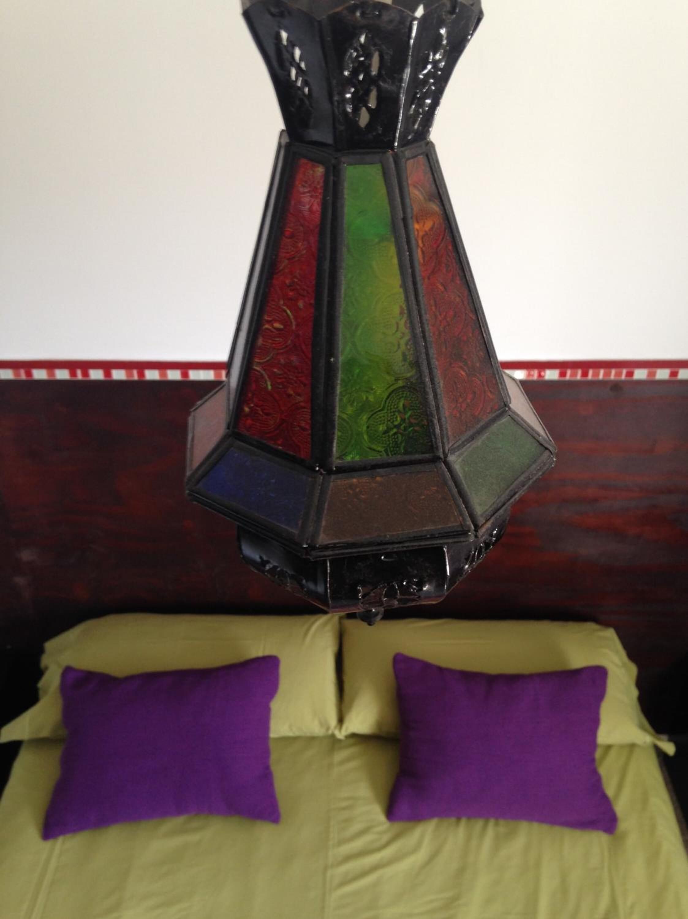 Una cama con sábanas verdes y almohadas moradas, un cabecero de madera oscura y una lámpara colgante negra y multicolora.