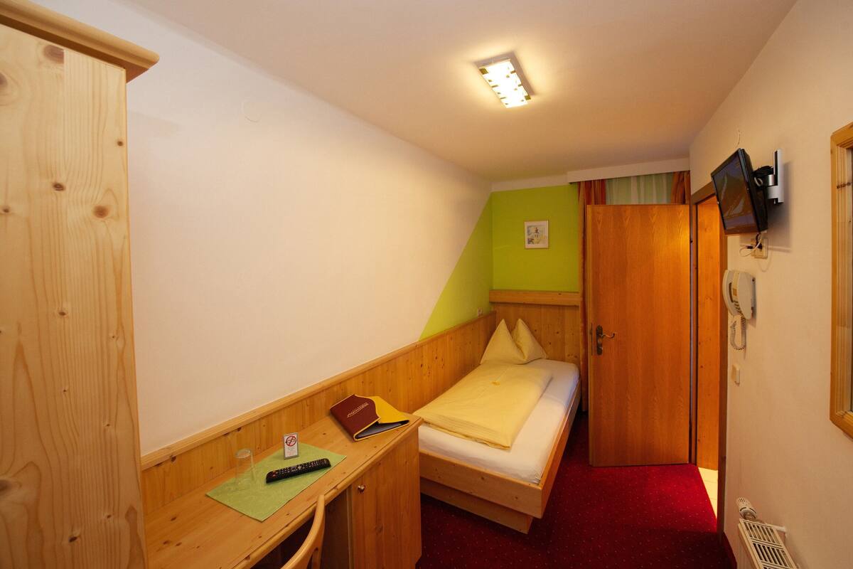 A small, compact room with a single bed, a desk, a wardrobe, and a television mounted on the wall.